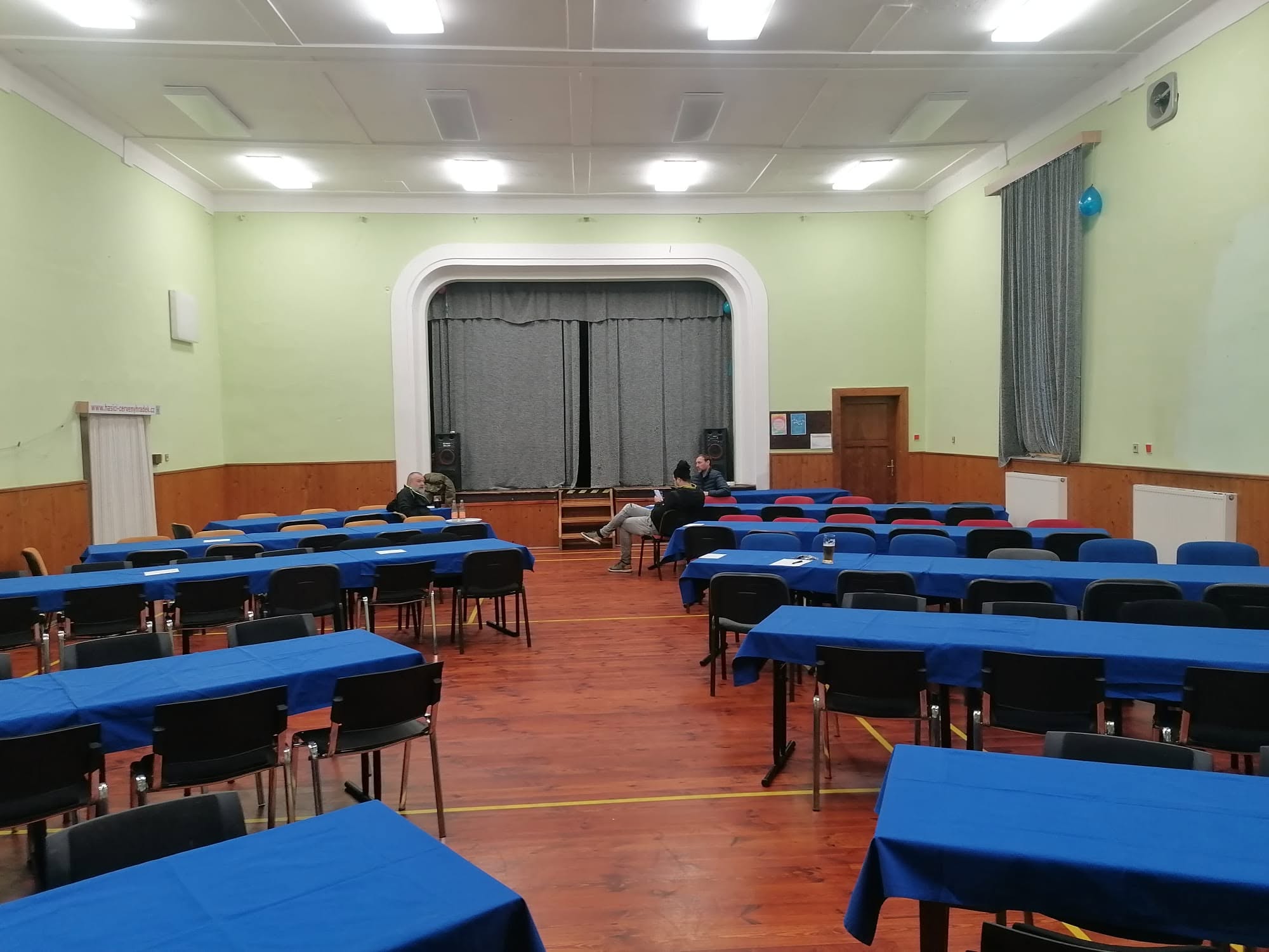 Sál se stoly s modrými ubrusy a jevištěm se zataženou oponou